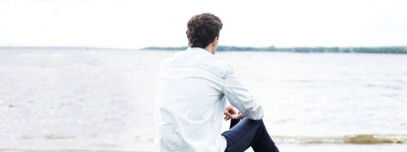 A young man sits on a beach and looks out at the water to represent the process of considering things before starting a business